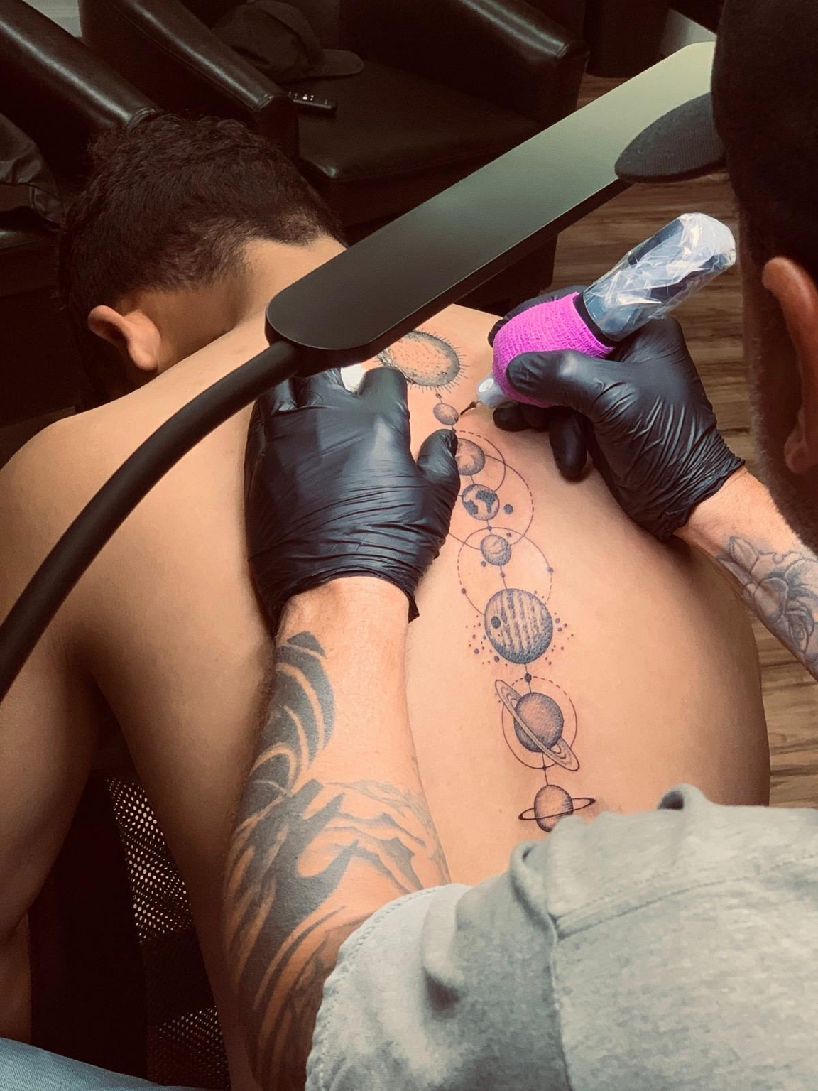 Nick tattooing a client's back with a tattoo machine while steadying the skin with his gloved hand.
